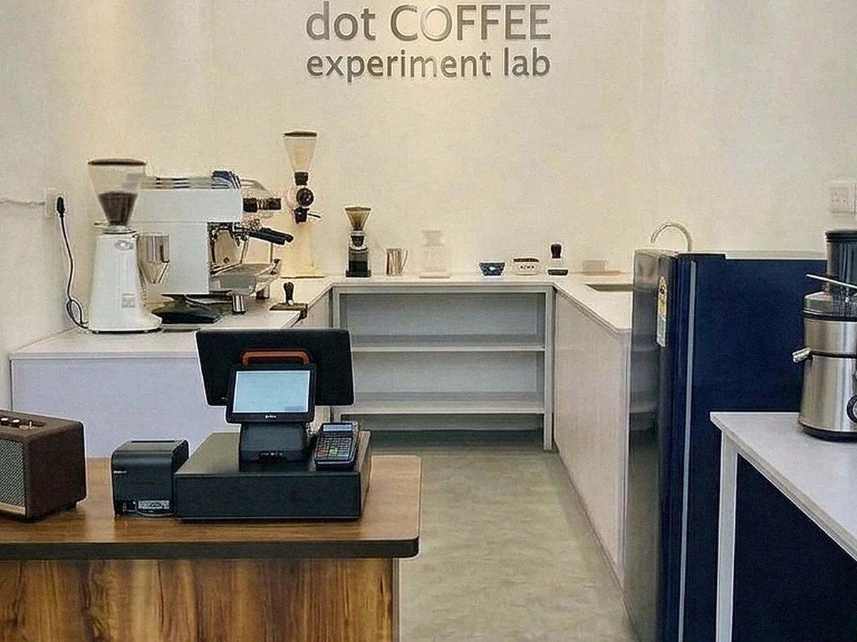 Inside the dot COFFEE experiment lab in Faridabad: espresso machine and grinders on a white counter, a wooden order counter with the till, and juicers on the right