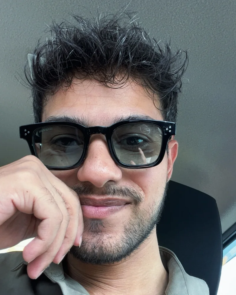 Aman smiling in a car selfie, wearing black-framed glasses, hand at his cheek